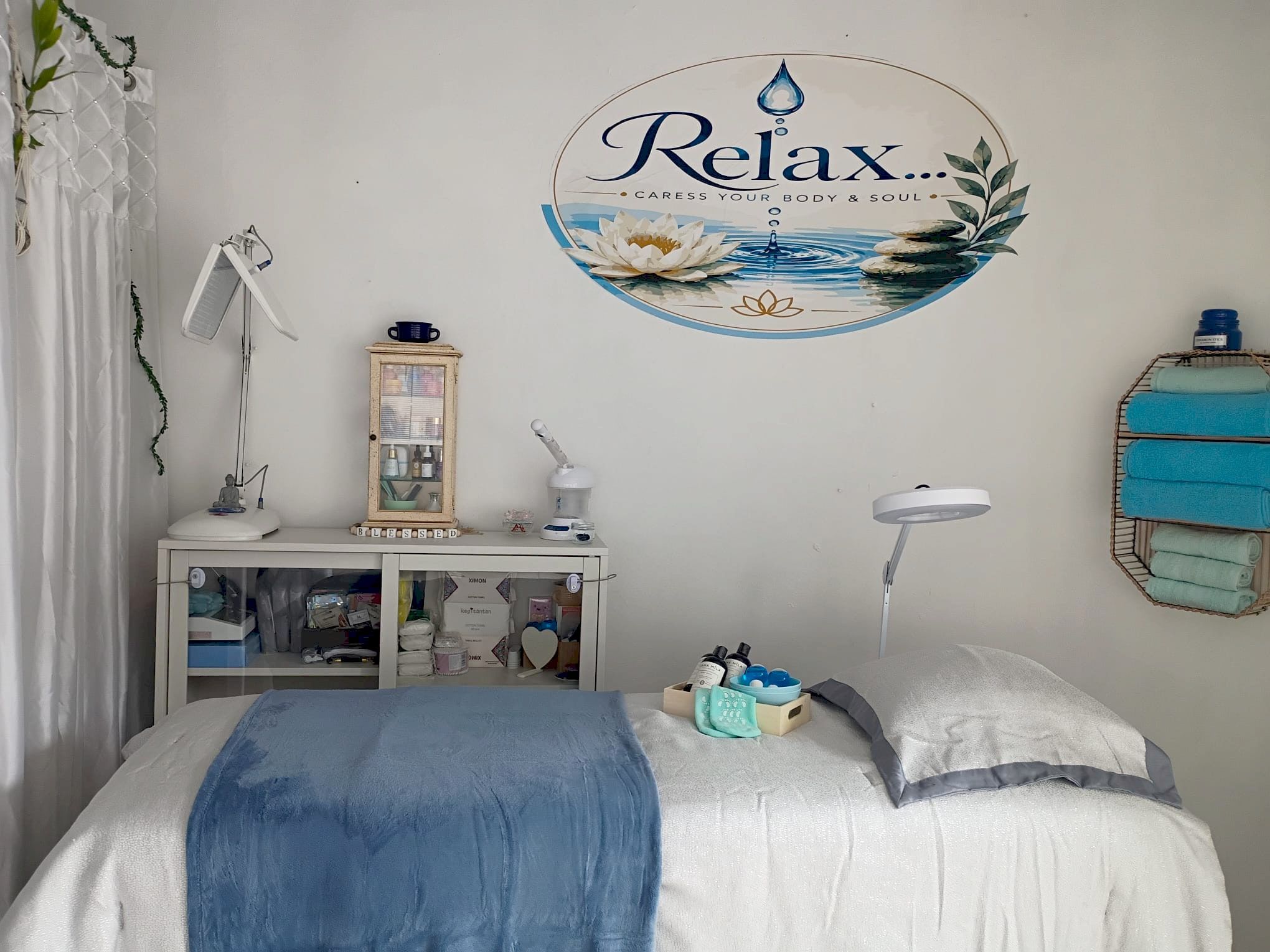Treatment room with the Relax logo on the wall, a bed with a blue blanket, a rack of turquoise towels and a cabinet of facial products.