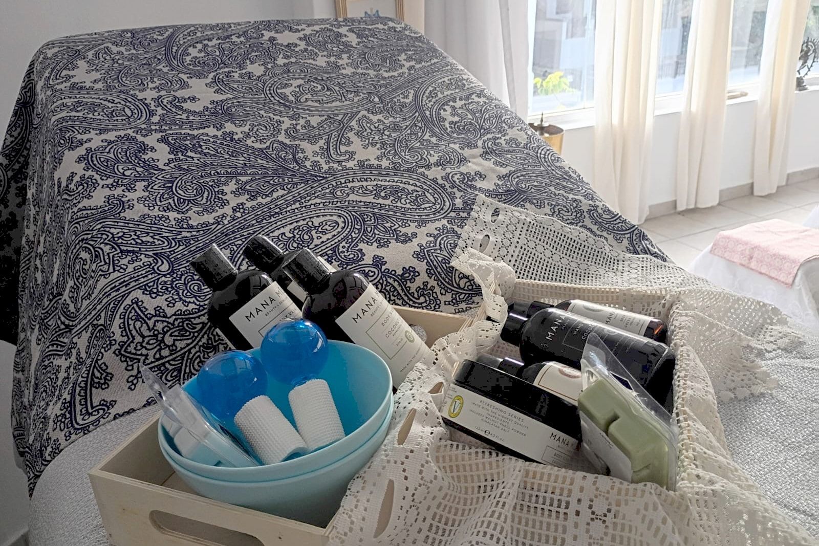 Treatment bed covered with a blue paisley throw, next to a tray of body-care products, facial ice globes and lace cloths, in a room with natural light.