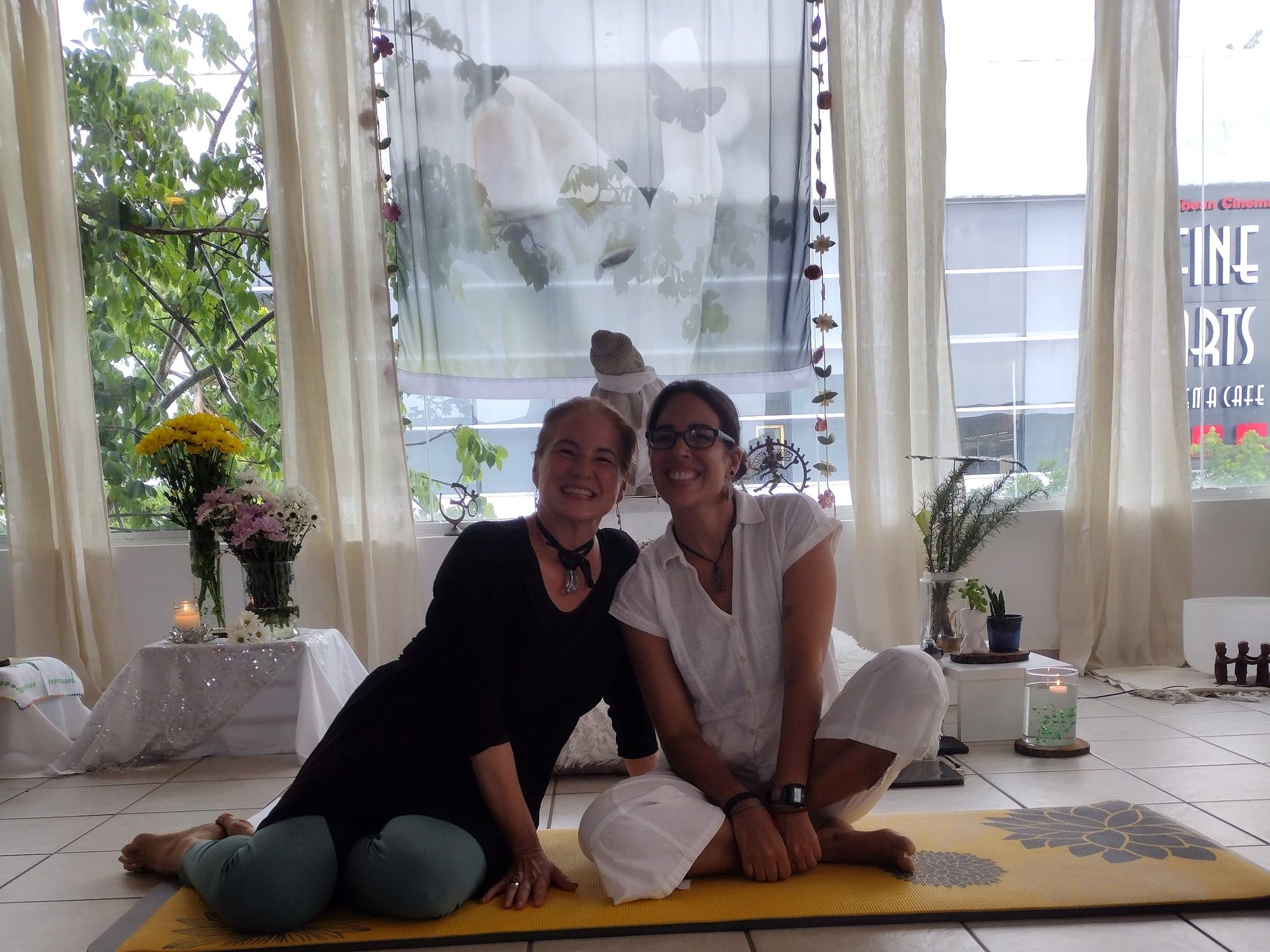 Two smiling women sitting together on a yellow mat, in front of windows with light curtains and flowers.