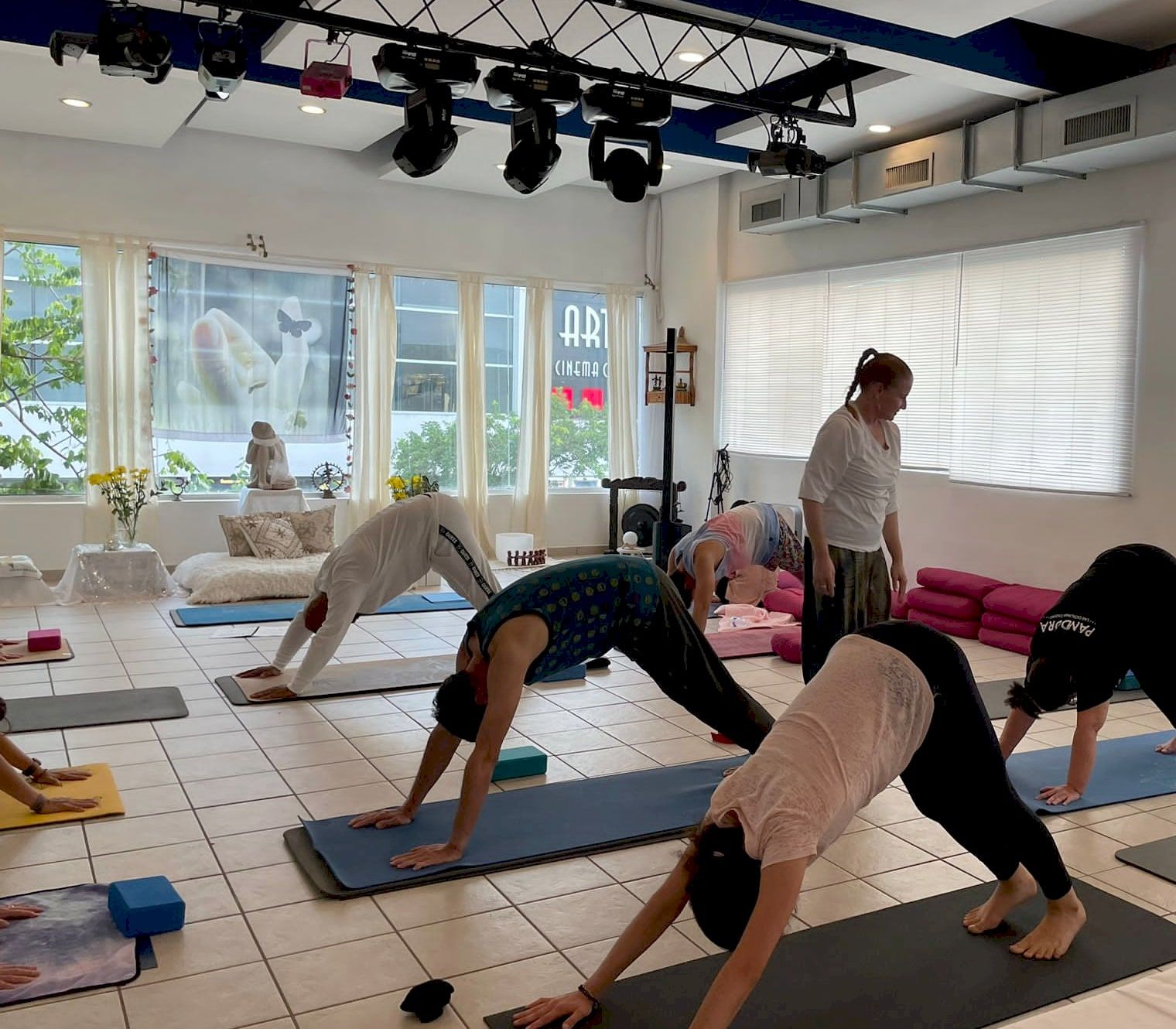 A group doing downward-facing dog on colorful mats in a bright room with large windows, while the teacher walks among them.