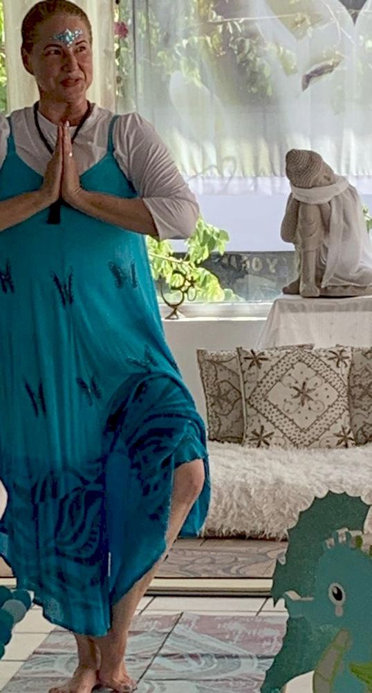A teacher in a turquoise dress in tree pose, palms together at her chest.